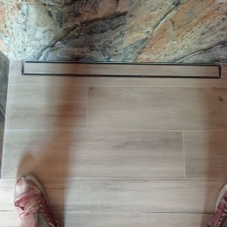 BUDO-FAST PROJECT - Ułożone jasne płytki podłogowe imitujące drewno z liniowym odpływem prysznicowym i fragmentem marmurowej ściany w tle, widoczne buty w dolnych rogach kadru.