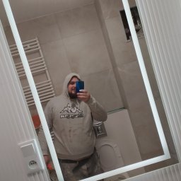 BUDO-FAST PROJECT - Selfie w lustrze łazienkowym: Osoba w bluzie z kapturem robi zdjęcie telefonem na tle wykończonej łazienki z płytkami i grzejnikiem drabinkowym.