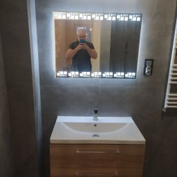 BUDO-FAST PROJECT - Wykończona łazienka w Zgierzu z umywalką na szafce z drewnopodobnym frontem i podświetlanym lustrem z geometrycznym wzorem, widoczne szare płytki na ścianach.