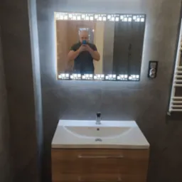 Wykończona łazienka w Zgierzu z umywalką na szafce z drewnopodobnym frontem i podświetlanym lustrem z geometrycznym wzorem, widoczne szare płytki na ścianach.