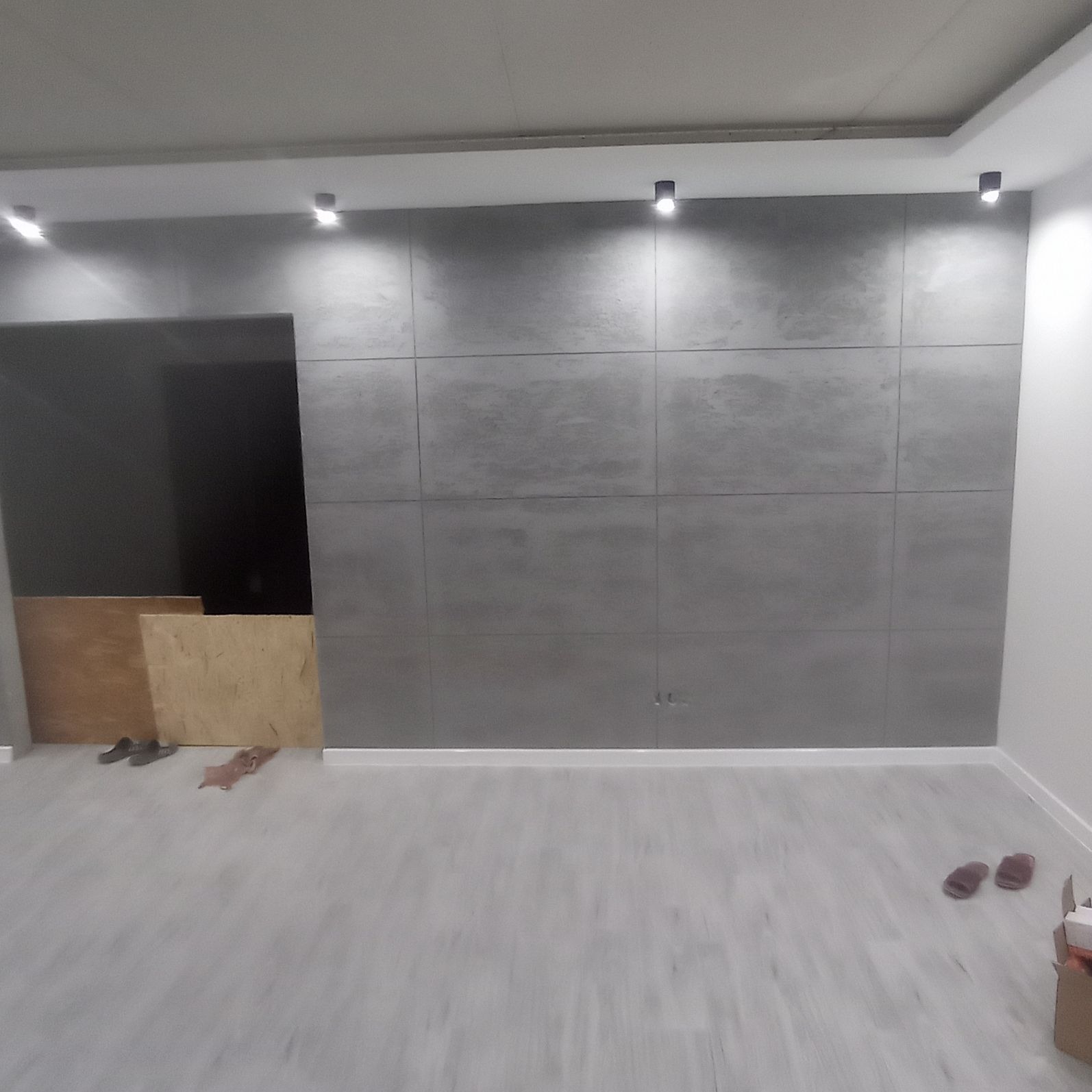 Szara ściana wykończona płytami imitującymi beton, z widocznymi punktowymi lampami sufitowymi i fragmentem niezabezpieczonej ściany z płytami OSB, podłoga wykończona panelami.