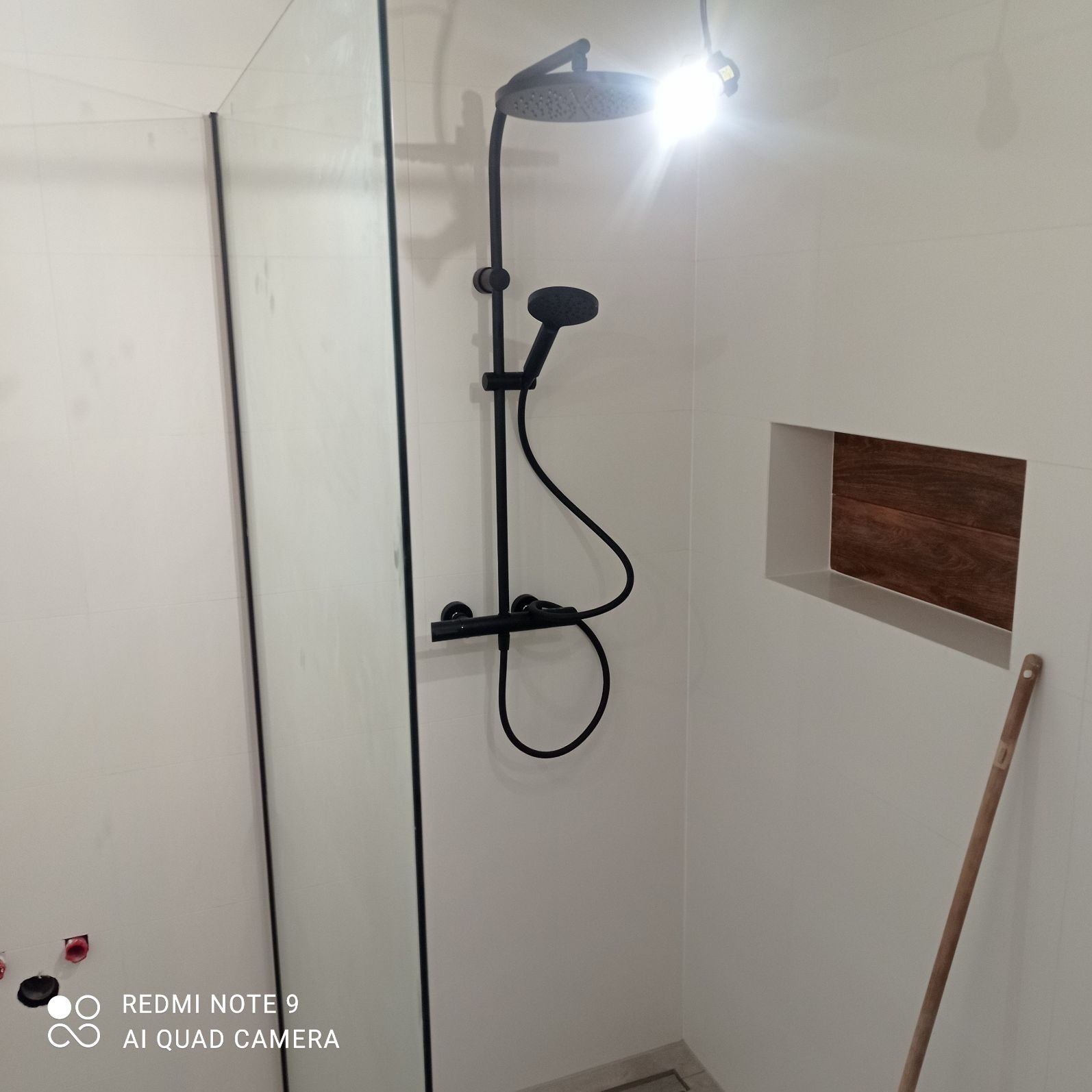 Nowoczesna kabina prysznicowa z czarną armaturą, szklaną ścianką i wnęką z drewnianym wykończeniem na ścianie pokrytej białymi płytkami.