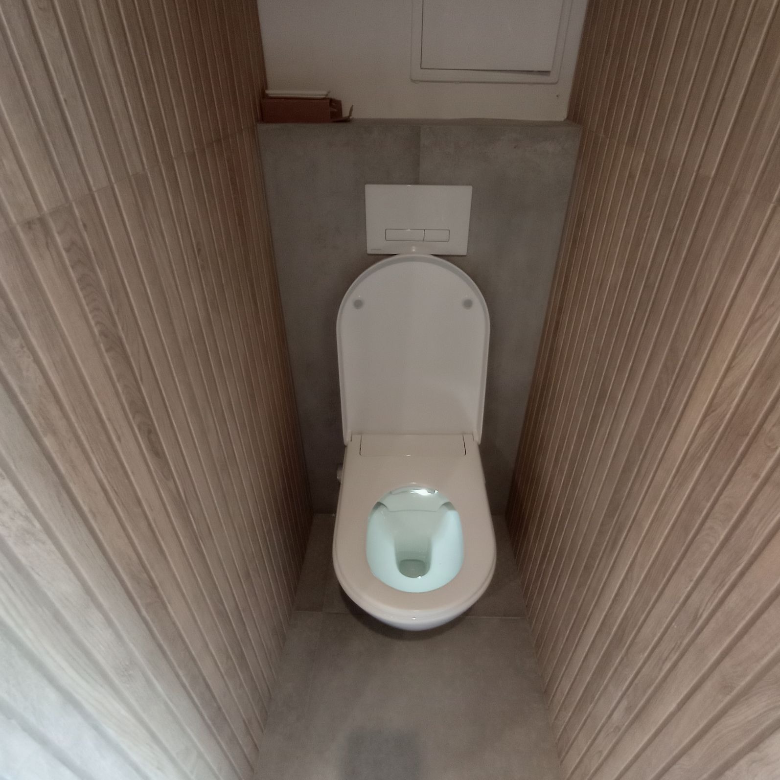 Wisząca toaleta z deską sedesową w łazience wykończonej panelami ściennymi imitującymi drewno, szara ściana za toaletą, widok z góry.