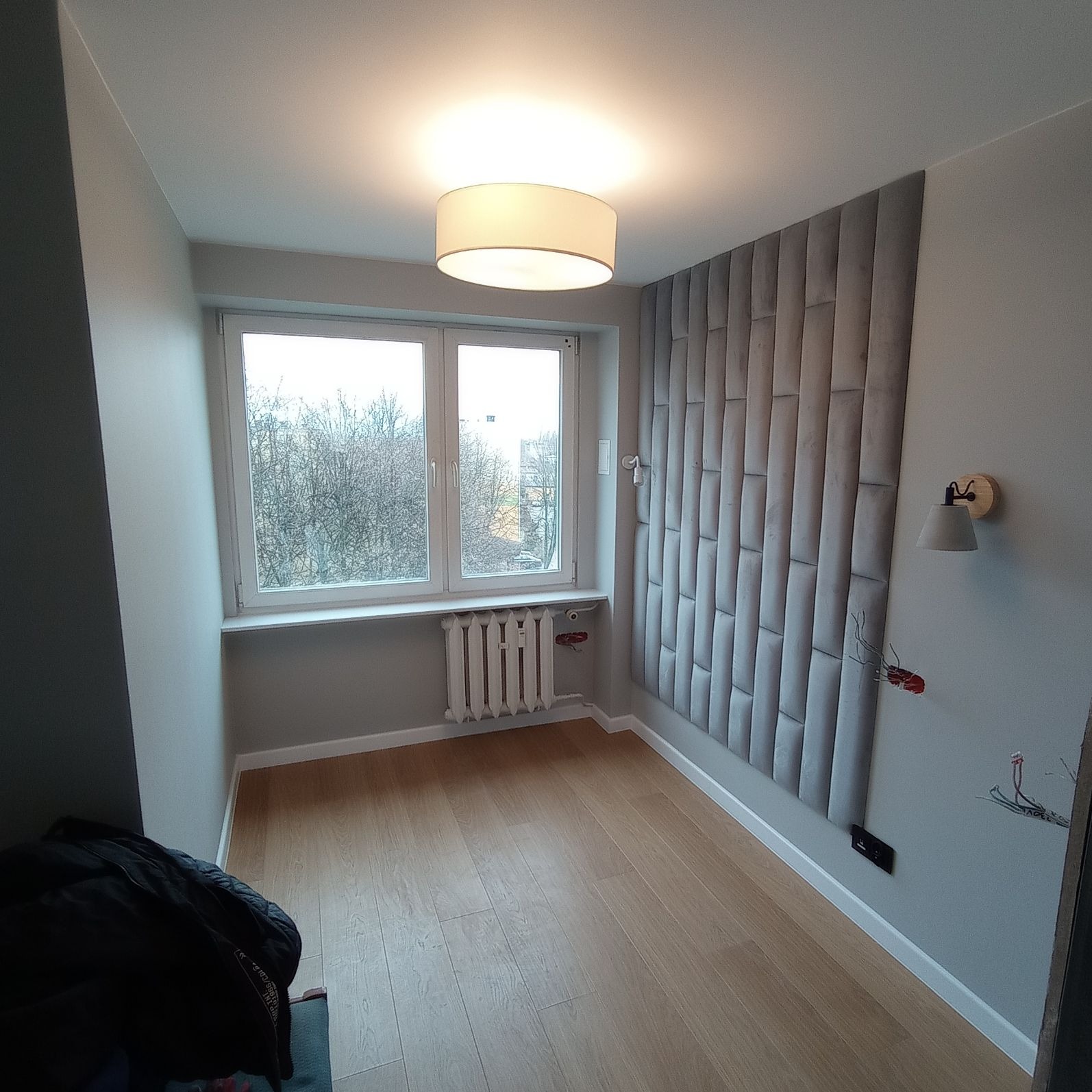 Pokój po remoncie: panele podłogowe w odcieniach jasnego drewna, szara ściana z panelami tapicerowanymi, białe listwy przypodłogowe, grzejnik pod oknem, widoczne kable elektryczne.