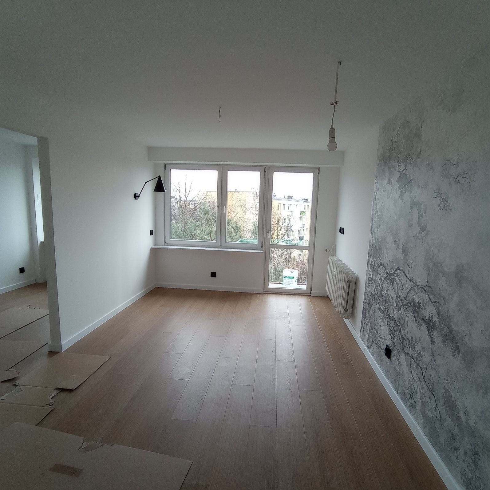 Pusty pokój po remoncie z drewnianą podłogą, świeżo pomalowanymi białymi ścianami i dekoracyjną ścianą z motywem drzewa, widok na okno z grzejnikiem i wiszącą żarówką.