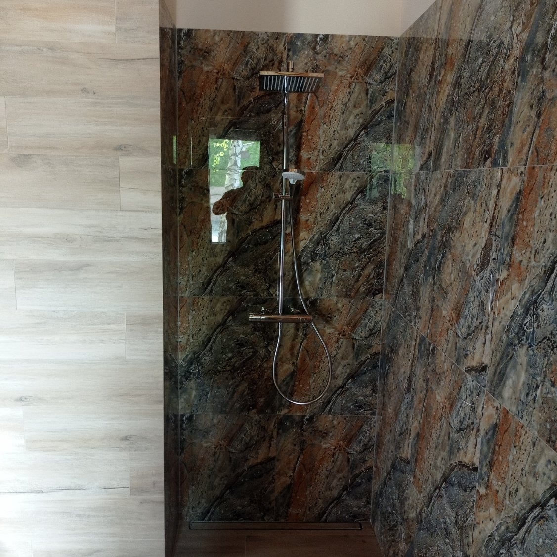 Wnętrze kabiny prysznicowej z marmurowymi płytkami w odcieniach brązu i szarości, kontrastujące z jasnymi, drewnopodobnymi płytkami na bocznej ścianie. Widoczna chromowana bateria i deszczownica.