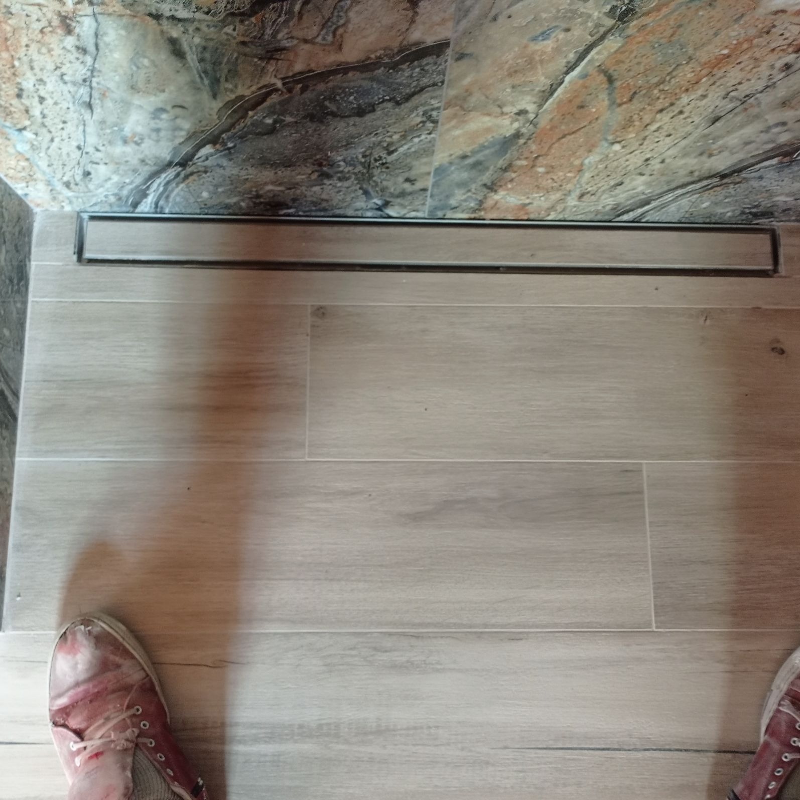 Ułożone jasne płytki podłogowe imitujące drewno z liniowym odpływem prysznicowym i fragmentem marmurowej ściany w tle, widoczne buty w dolnych rogach kadru.