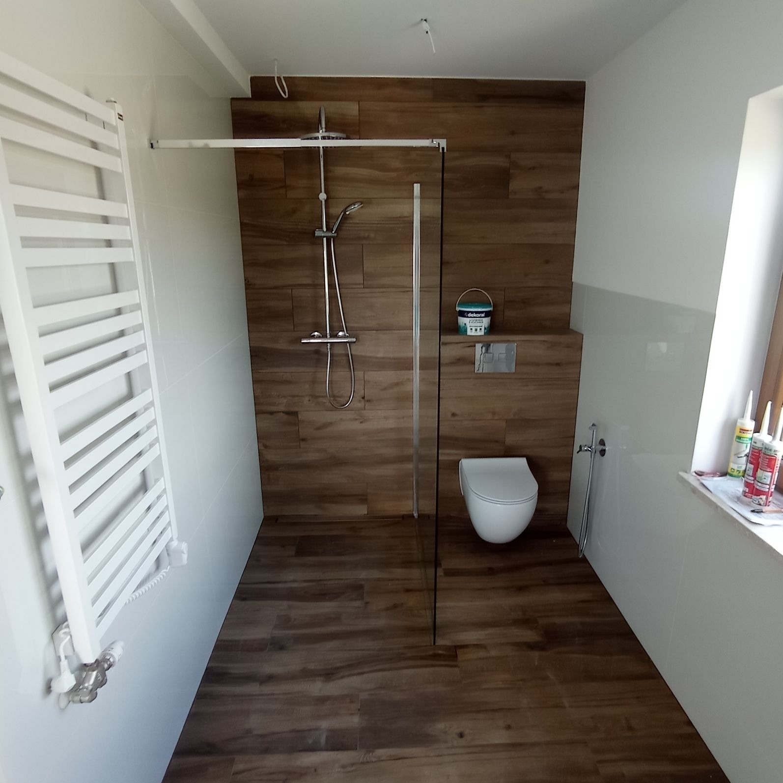Nowoczesna łazienka z drewnopodobnymi płytkami, kabiną prysznicową typu walk-in, wiszącą toaletą i grzejnikiem drabinkowym, widoczny pojemnik z farbą 'Dekoral'
