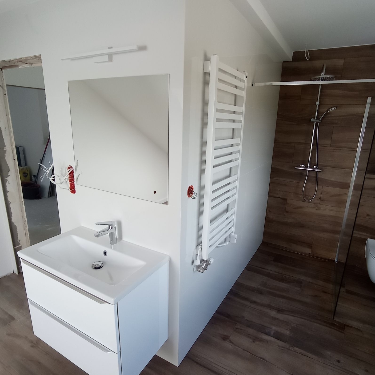 Biała umywalka z szafką, lustro z podświetleniem, grzejnik drabinkowy i kabina prysznicowa z panelami imitującymi drewno w trakcie wykańczania łazienki, widoczne niedokończone ściany i przewody...