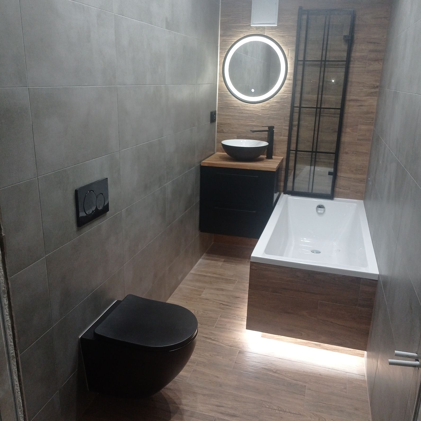 Nowoczesna łazienka z szarymi płytkami, podwieszaną czarną toaletą, umywalką w kształcie misy na drewnianym blacie, okrągłym lustrem z podświetleniem i kabiną prysznicową z czarną ramą.