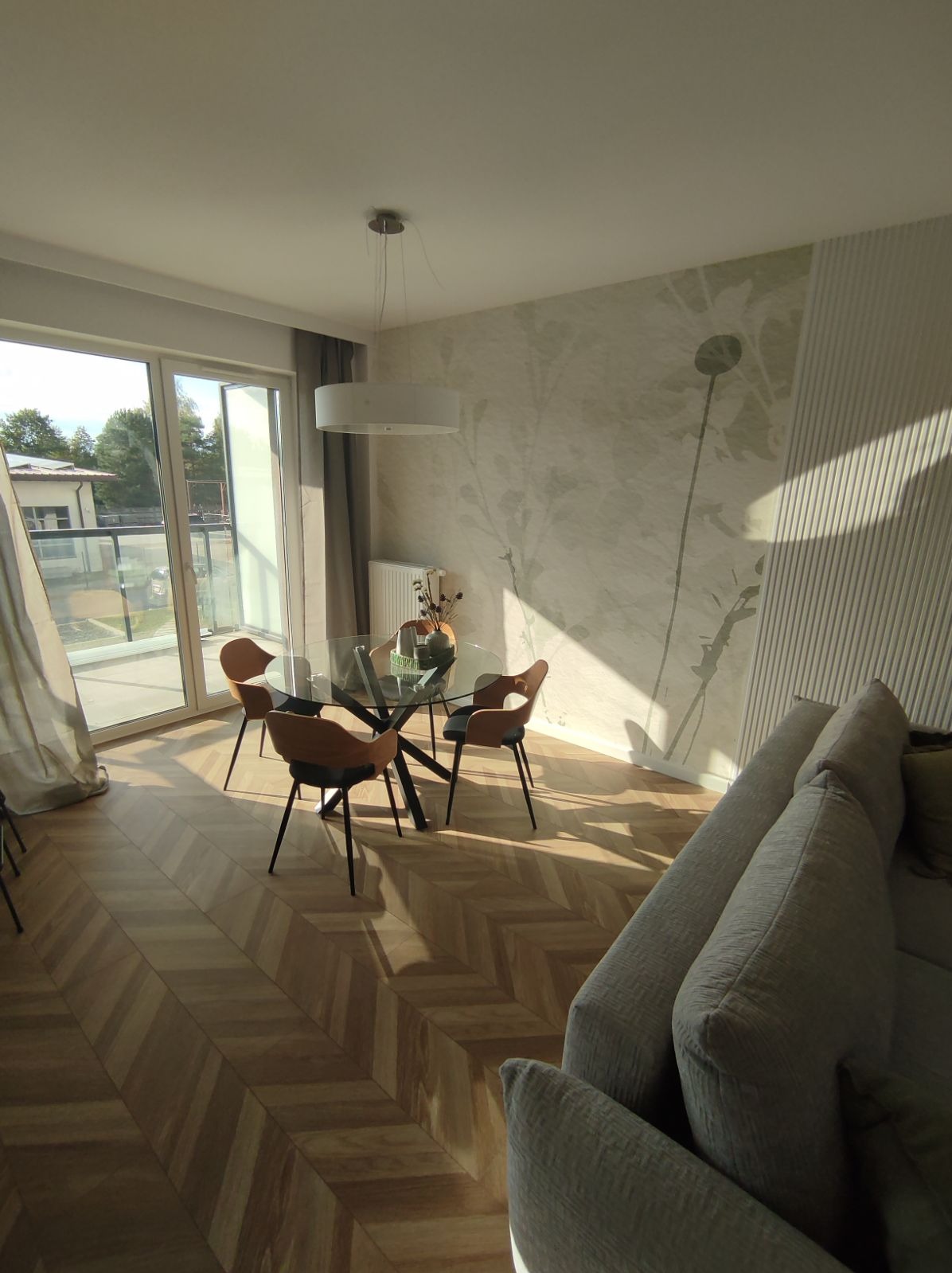 Jasne wnętrze salonu z parkietem ułożonym w jodełkę, szklanym stołem z czterema krzesłami, tapetą w kwiatowe wzory i fragmentem szarej sofy, oświetlone popołudniowym słońcem wpadającym przez okno...