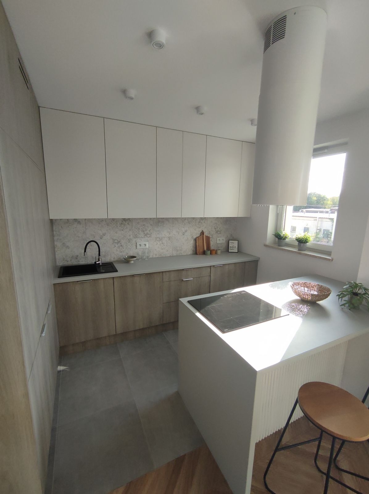 Nowoczesna kuchnia z białymi szafkami górnymi i drewnopodobnymi dolnymi, wyspą z płytą indukcyjną, okrągłym okapem i szarymi płytkami na ścianie.