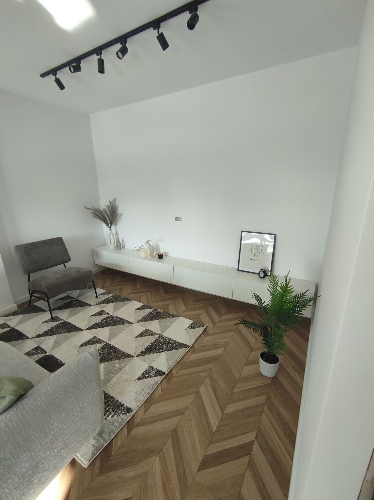 Jasne wnętrze salonu z parkietem ułożonym we wzór jodełki, szarym fotelem, dywanem w geometryczne wzory i białą szafką RTV z dekoracjami.