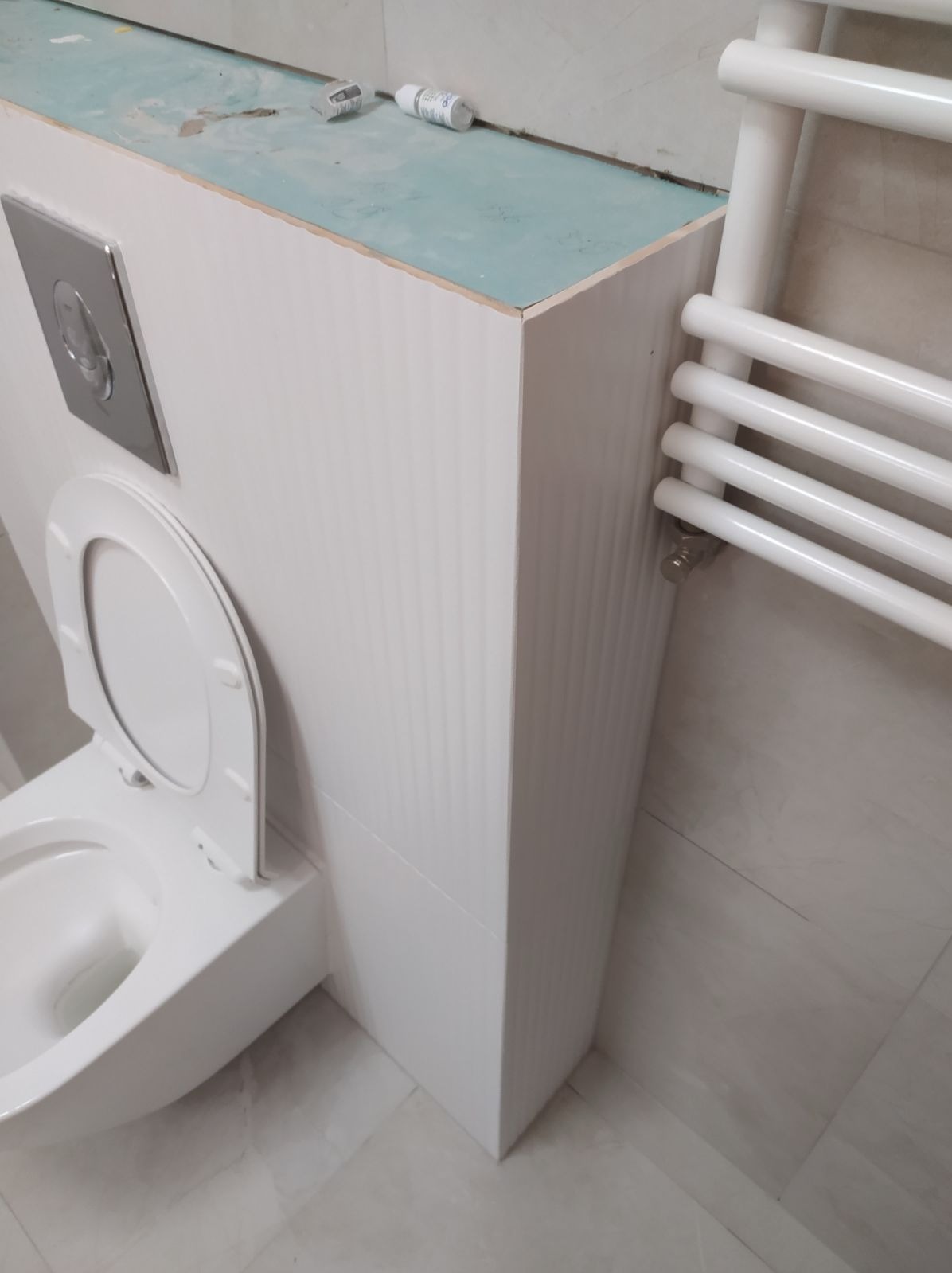 Biała toaleta podwieszana z otwartą deską, zabudowana białą płytą z widocznym stelażem i przyciskiem spłukiwania, obok grzejnik drabinkowy, widoczne ślady prac wykończeniowych na górnej krawędzi...