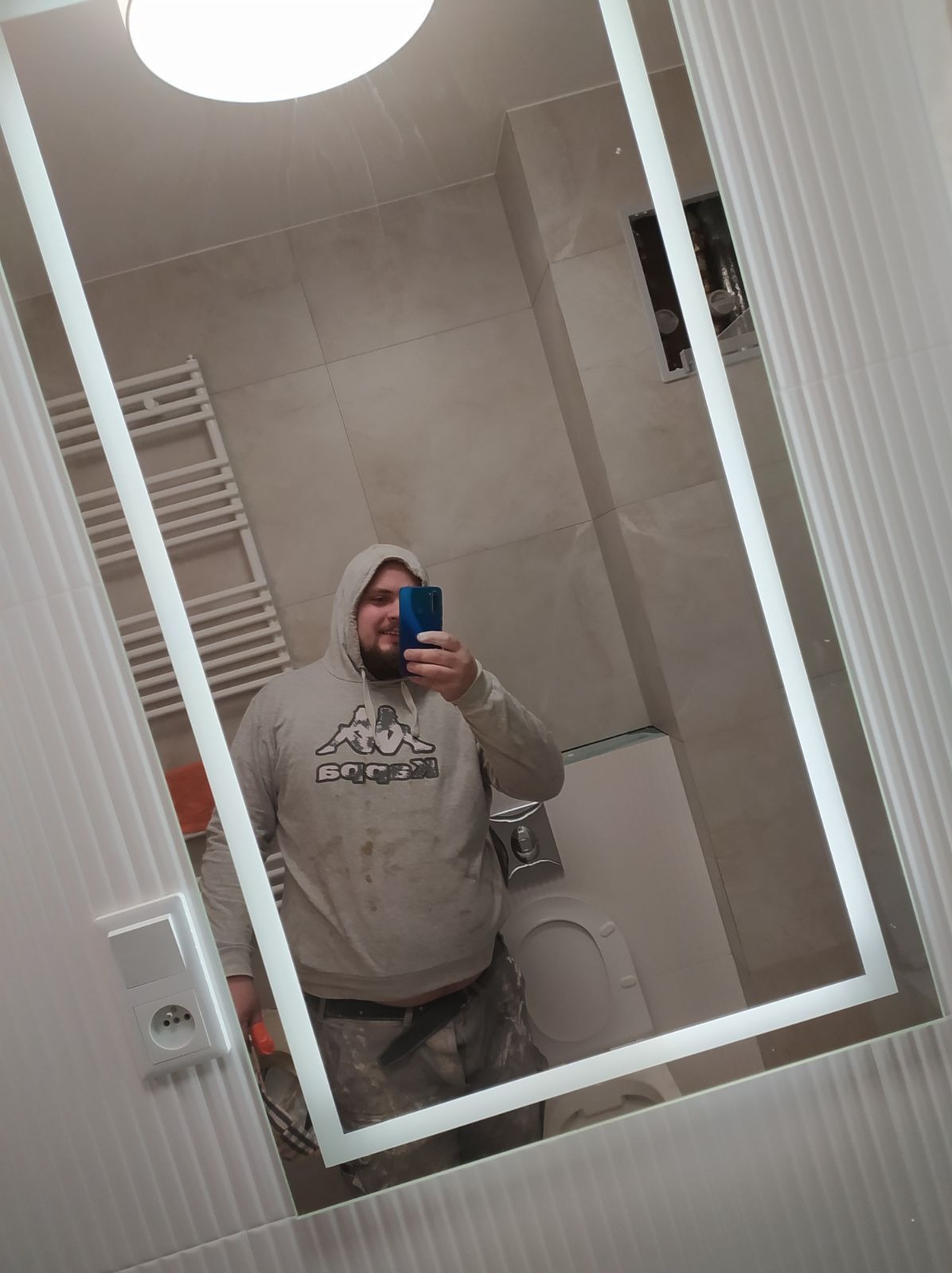 Selfie w lustrze łazienkowym: Osoba w bluzie z kapturem robi zdjęcie telefonem na tle wykończonej łazienki z płytkami i grzejnikiem drabinkowym.