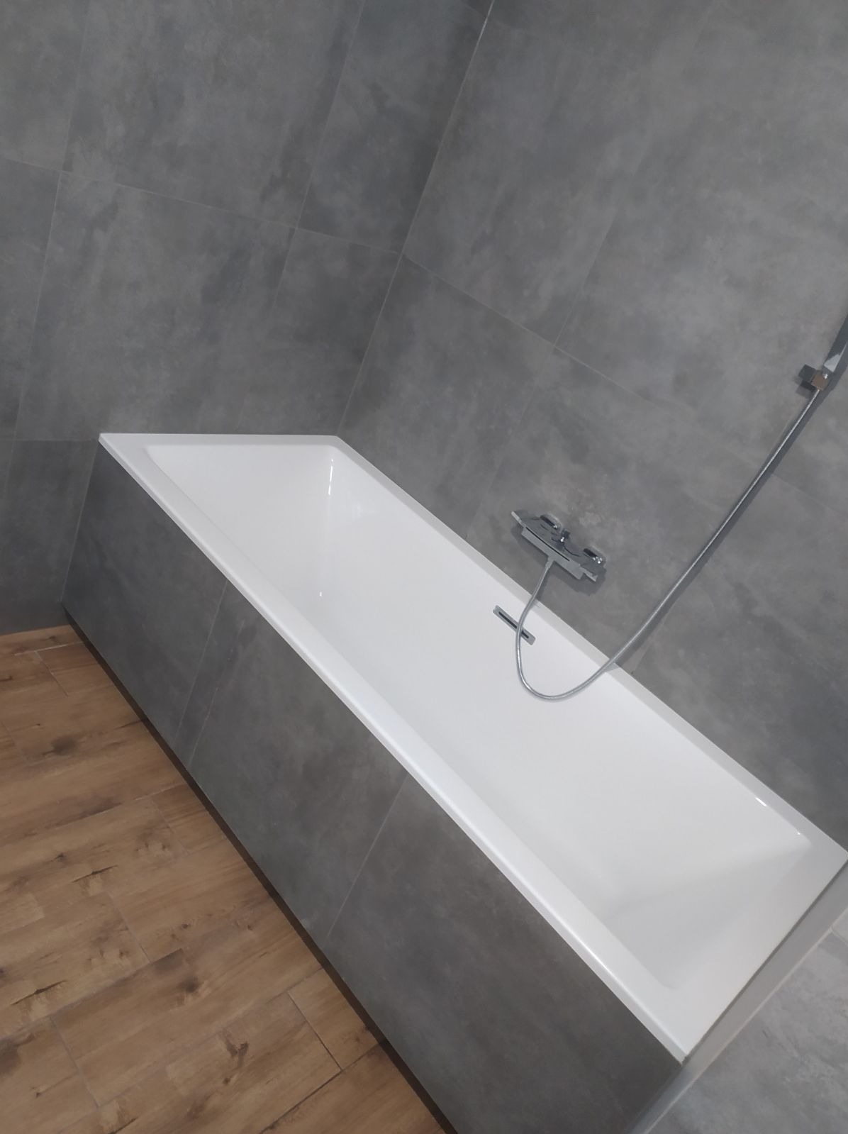 Nowoczesna biała wanna obudowana szarymi płytkami, z chromowaną baterią i prysznicem, na podłodze panele imitujące drewno, szare płytki na ścianach.