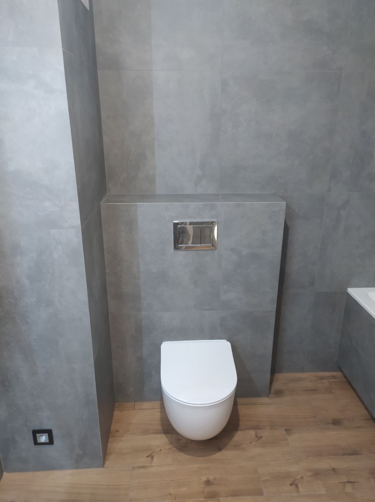 Nowoczesna toaleta podwieszana z chromowanym przyciskiem spłukującym na tle szarych, matowych płytek imitujących beton i drewnopodobnej podłogi.
