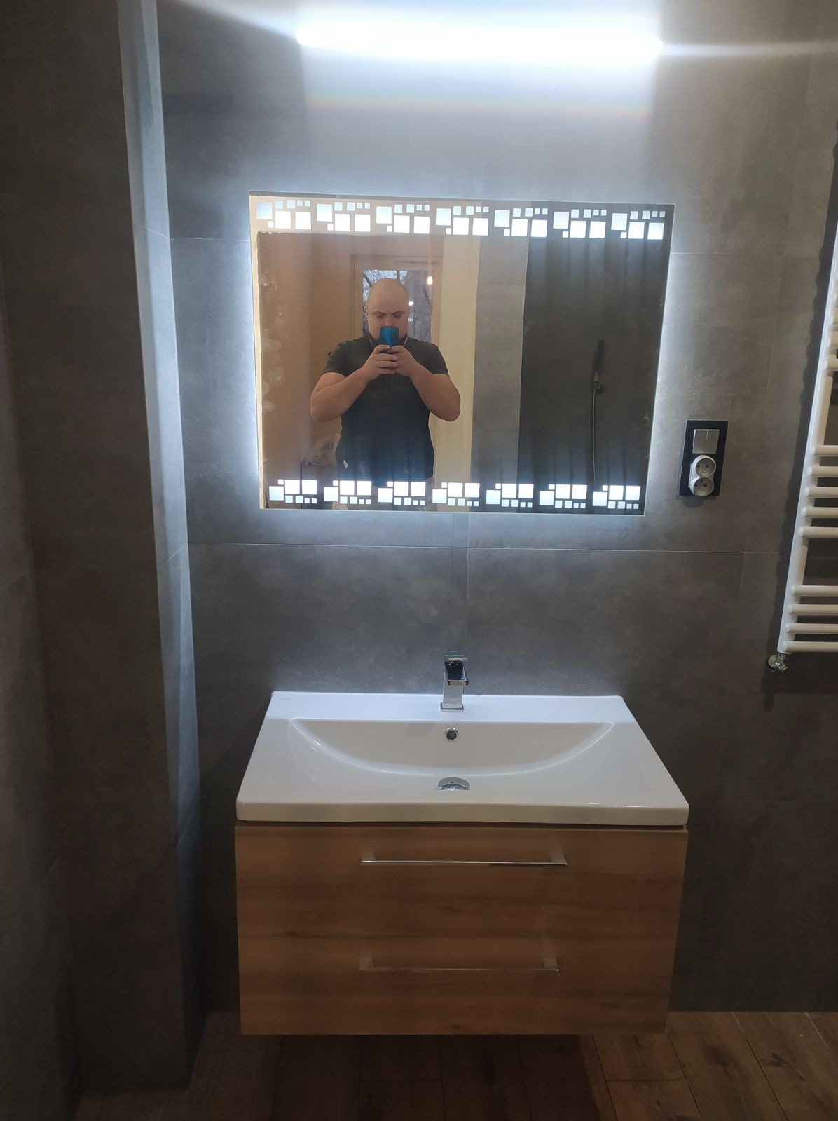 Wykończona łazienka w Zgierzu z umywalką na szafce z drewnopodobnym frontem i podświetlanym lustrem z geometrycznym wzorem, widoczne szare płytki na ścianach.