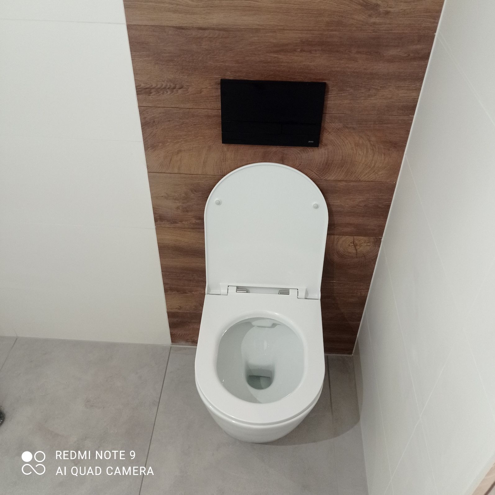 Nowoczesna toaleta podwieszana z czarnym przyciskiem spłukującym na tle paneli imitujących drewno, białe płytki na ścianach i szara podłoga.