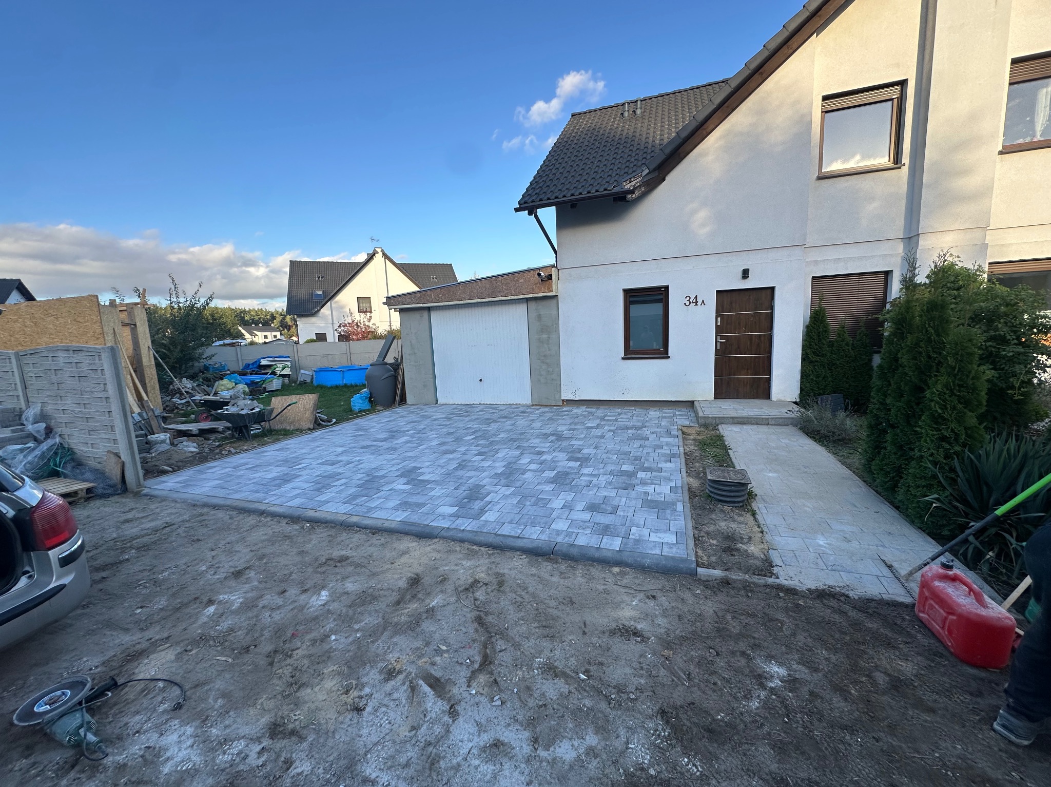 Ułożona szara kostka brukowa na podjeździe w Lesznie, widoczne narzędzia i częściowo rozkopany teren, dom z numerem 34A w tle.