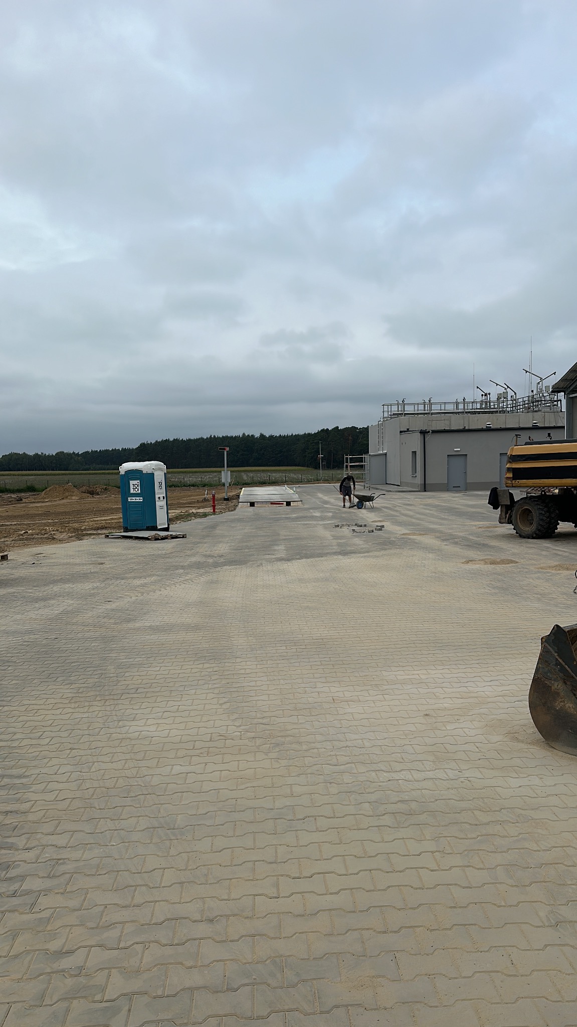 Rozległy plac wyłożony szarą kostką brukową typu behaton, z widocznym pracownikiem z taczką w oddali, przenośną toaletą TOI TOI, budynkiem przemysłowym i fragmentem ciężkiego sprzętu po prawej...