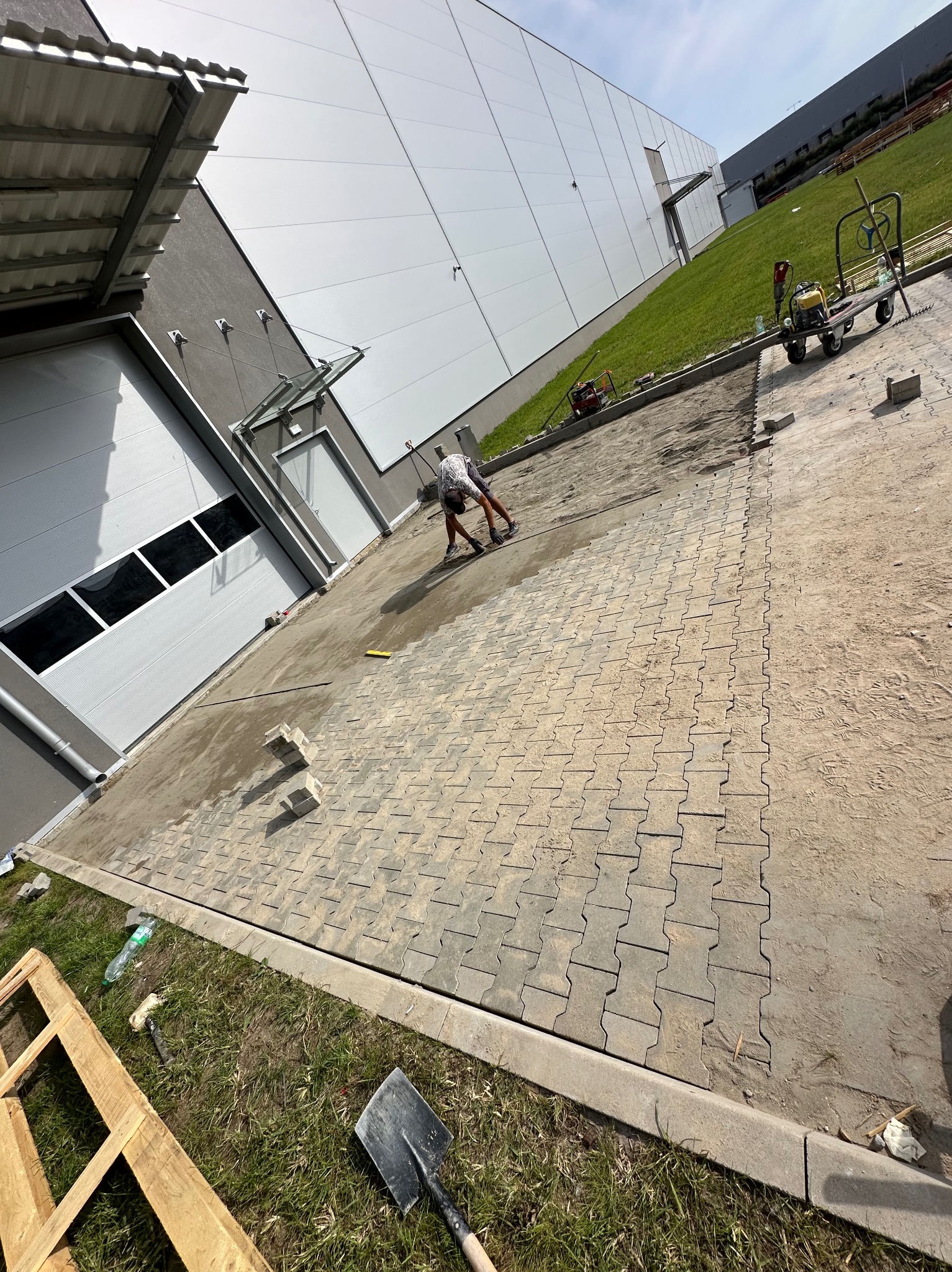 Pracownik w trakcie układania kostki brukowej typu 'H' na podjeździe przed budynkiem przemysłowym, widoczne narzędzia i materiały budowlane.