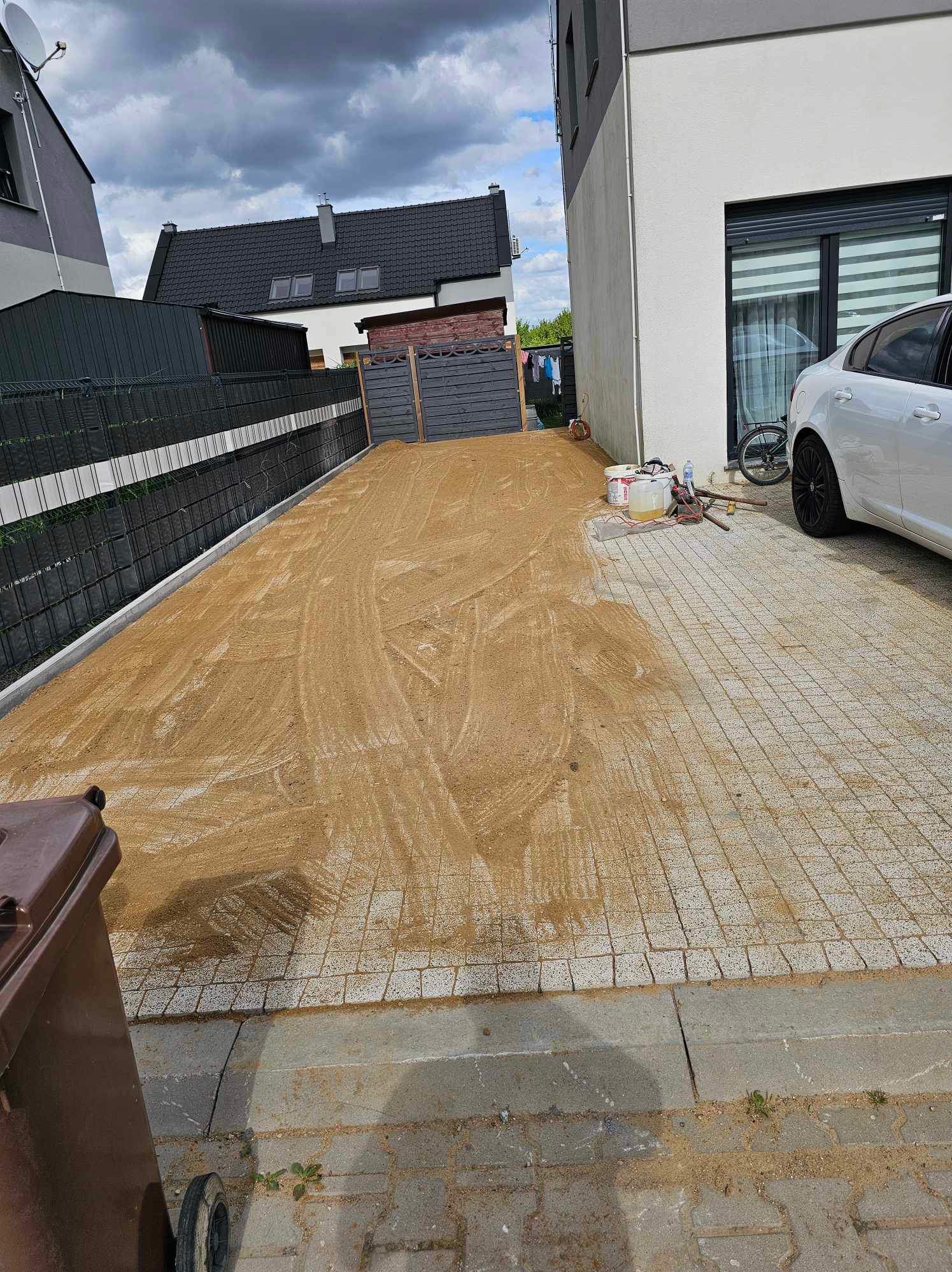 Przygotowanie podłoża pod nawierzchnię z kostki: wyrównany piasek na ułożonej siatce z kostki, widoczne narzędzia i pojemniki z zaprawą, w tle dom jednorodzinny i zaparkowany samochód.