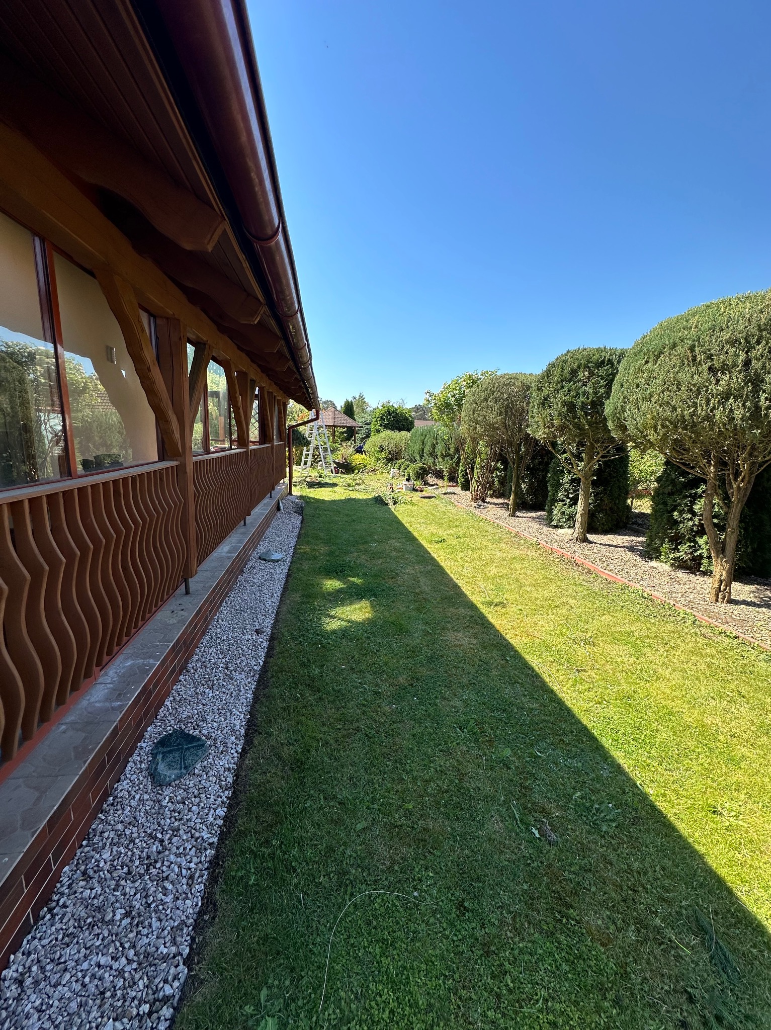 Widok na zadbany ogród z równo przystrzyżonym trawnikiem, szpalerem formowanych drzew i drewnianą altaną w tle, oświetlony jasnym światłem słonecznym.