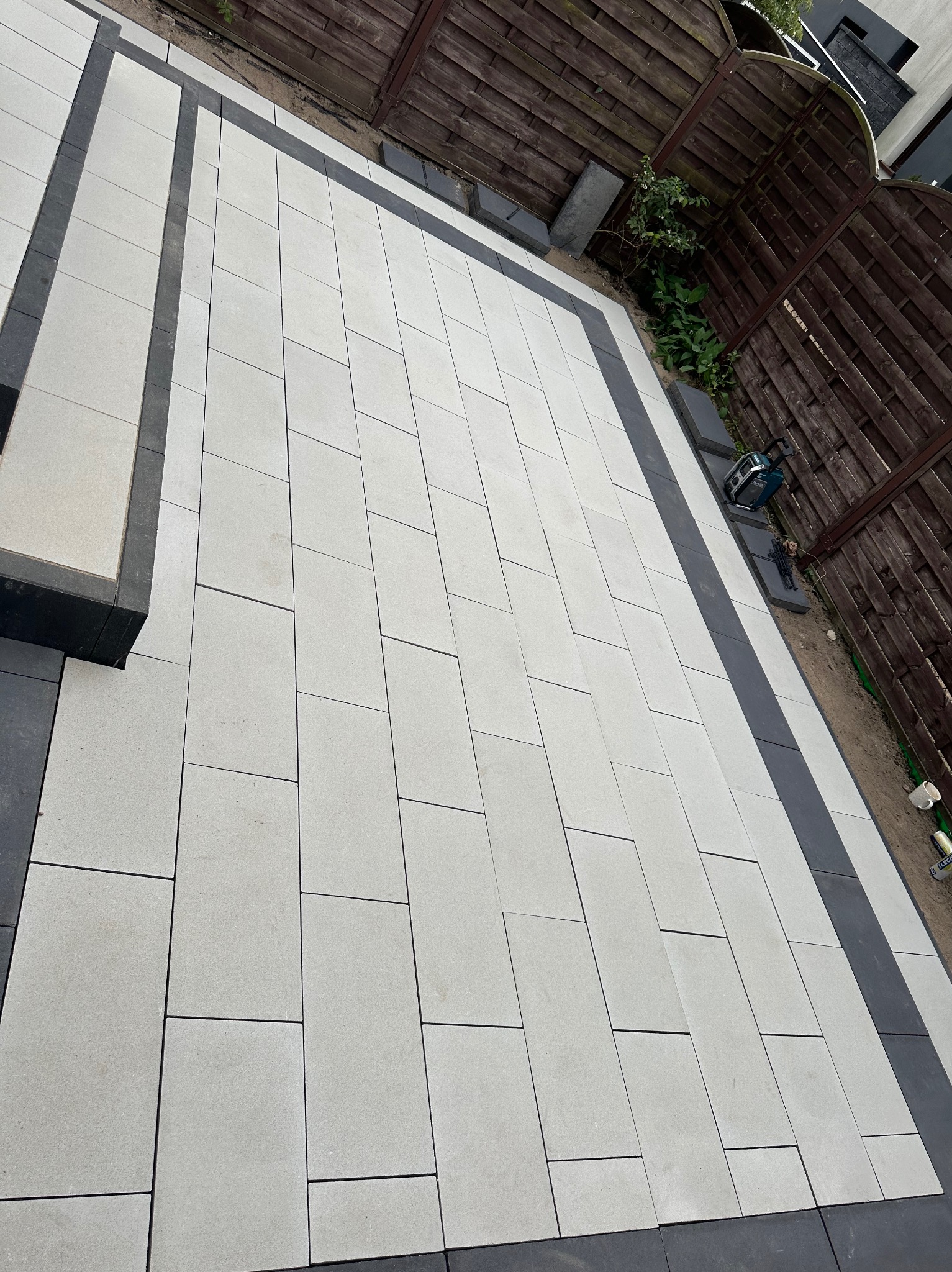 Nowo ułożony taras z jasnoszarych prostokątnych płyt betonowych z ciemnoszarym obramowaniem, widoczna poziomica na boku tarasu, brązowe ogrodzenie w tle.