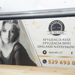 PRZEDSIĘBIORSTWO USŁUGOWO- HANDLOWE SERHII LUHOVYI - Wydrukowany baner reklamowy salonu kosmetycznego 'Sunlash' z czarno-białym portretem kobiety, informacjami kontaktowymi i ofertą stylizacji rzęs, brwi i opalania natryskowego, umieszczony przed...