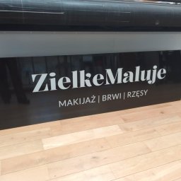 PRZEDSIĘBIORSTWO USŁUGOWO- HANDLOWE SERHII LUHOVYI - Czarna tablica z białym napisem 'ZielkeMaluje. Makijaż | Brwi | Rzęsy' ustawiona na drewnianej podłodze, w tle widoczna drukarka wielkoformatowa.