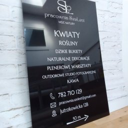 PRZEDSIĘBIORSTWO USŁUGOWO- HANDLOWE SERHII LUHOVYI - Czarny, błyszczący szyld reklamowy z białym napisem 'Pracownia SzeLest' i informacjami o kwiaciarni, roślinach, warsztatach plenerowych, studio fotograficznym, kawie oraz danymi kontaktowymi...