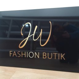 PRZEDSIĘBIORSTWO USŁUGOWO- HANDLOWE SERHII LUHOVYI - Czarna, błyszcząca tablica z napisem 'JW FASHION BUTIK' w kolorze złotym, ustawiona na drewnianej podłodze przy białej ścianie; widoczne odbicie osoby fotografującej.