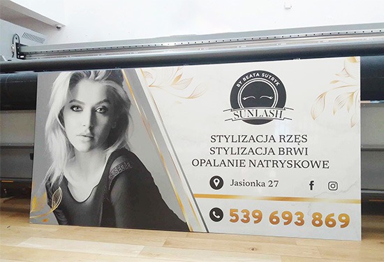 Wydrukowany baner reklamowy salonu kosmetycznego 'Sunlash' z czarno-białym portretem kobiety, informacjami kontaktowymi i ofertą stylizacji rzęs, brwi i opalania natryskowego, umieszczony przed...