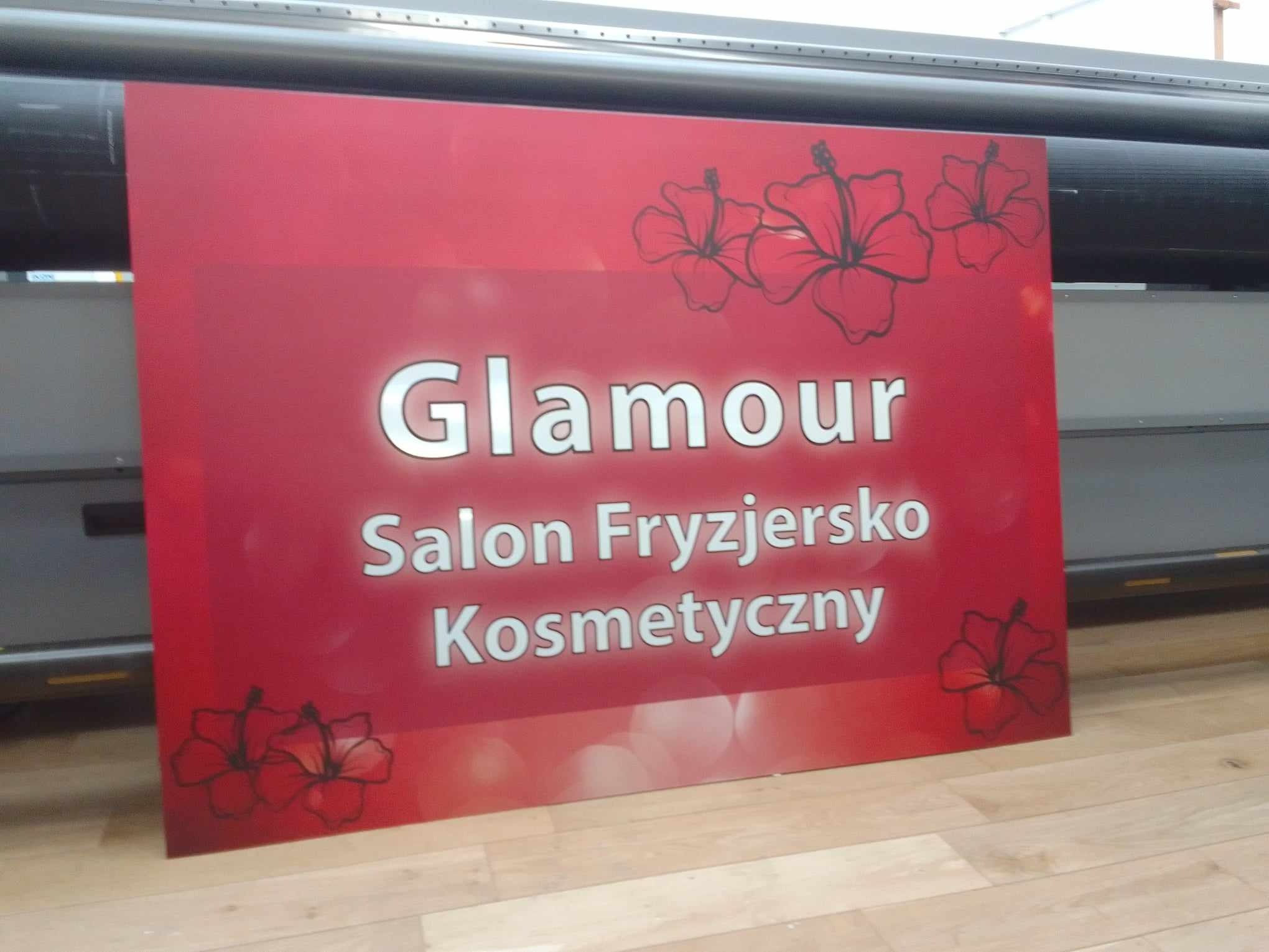 Czerwony szyld z napisem 'Glamour Salon Fryzjersko Kosmetyczny' i motywem kwiatowym, wyprodukowany we Wrocławiu techniką druku wielkoformatowego, widoczny na tle maszyny drukarskiej.