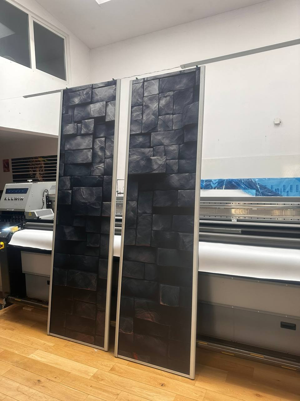 Dwa panele z nadrukiem imitującym mur z ciemnych, trójwymiarowych bloków, ustawione pionowo na podłodze z parkietu, w tle widoczna maszyna drukarska wielkoformatowa.