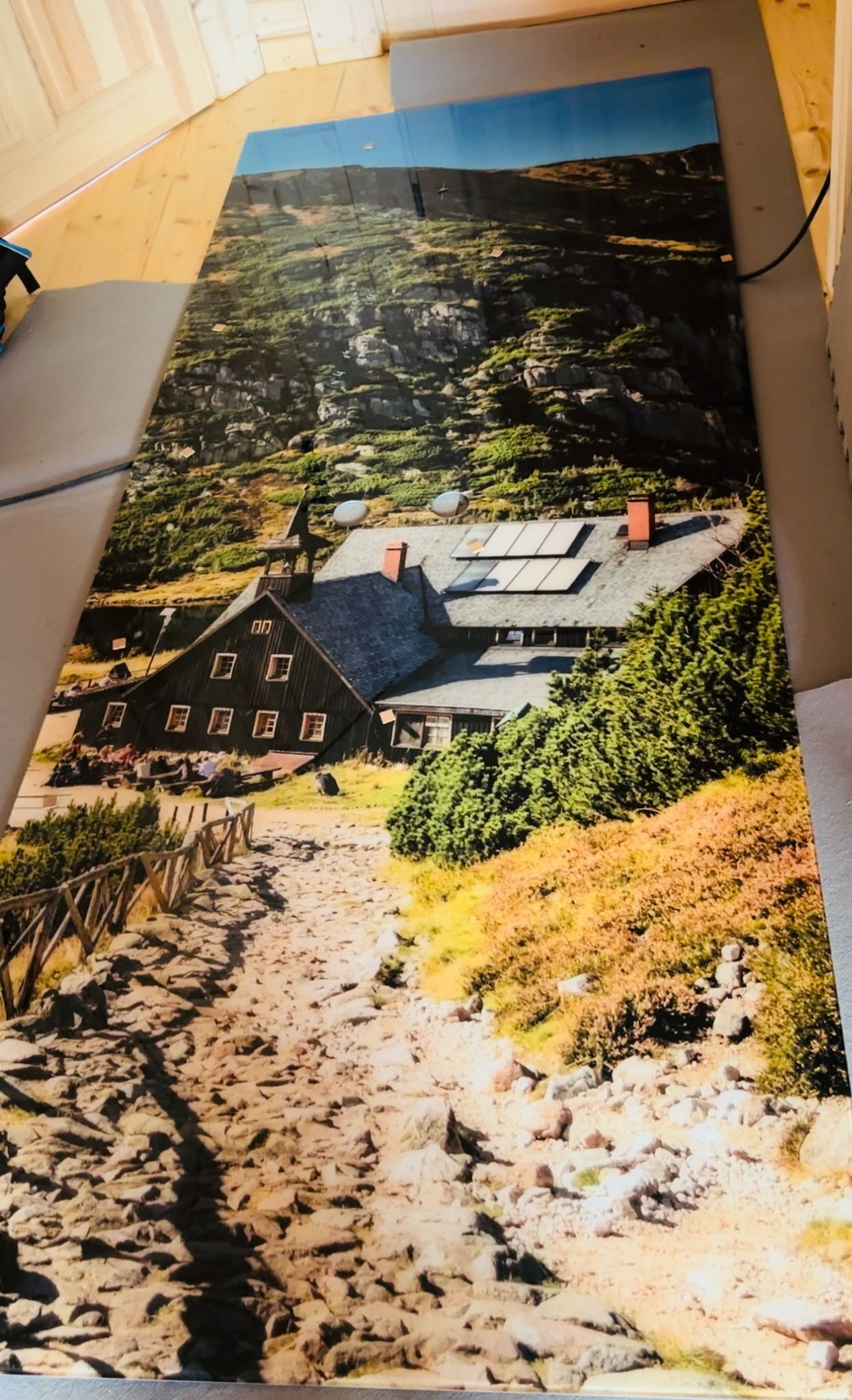 Duży wydruk przedstawiający górski krajobraz z drewnianym schroniskiem z czerwonymi dachami i kamienną ścieżką, widoczne panele słoneczne na dachu.