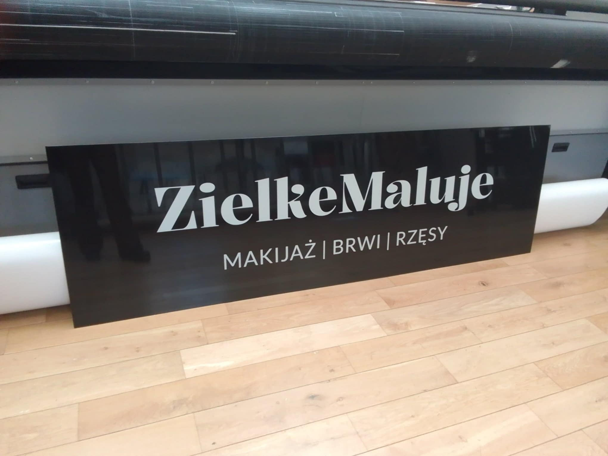 Czarna tablica z białym napisem 'ZielkeMaluje. Makijaż | Brwi | Rzęsy' ustawiona na drewnianej podłodze, w tle widoczna drukarka wielkoformatowa.