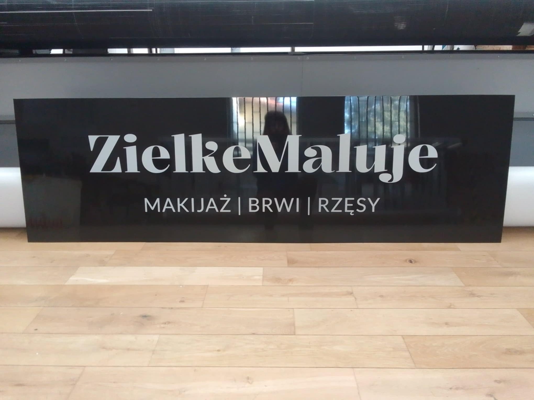 Czarny szyld z białym napisem 'ZielkeMaluje MAKIJAŻ | BRWI | RZĘSY' stojący na podłodze z paneli, widoczny w tle fragment drukarki.
