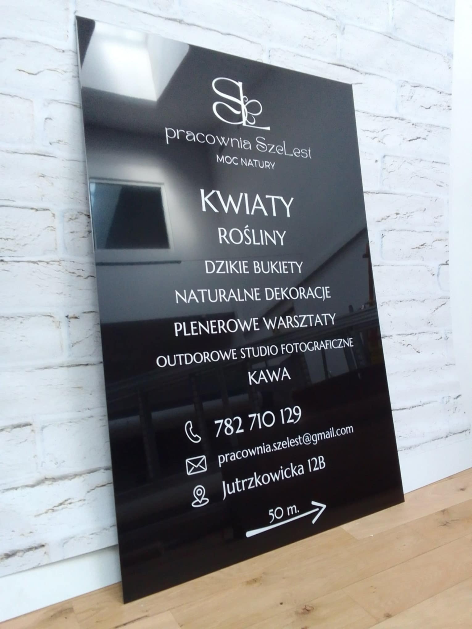 Czarny, błyszczący szyld reklamowy z białym napisem 'Pracownia SzeLest' i informacjami o kwiaciarni, roślinach, warsztatach plenerowych, studio fotograficznym, kawie oraz danymi kontaktowymi...