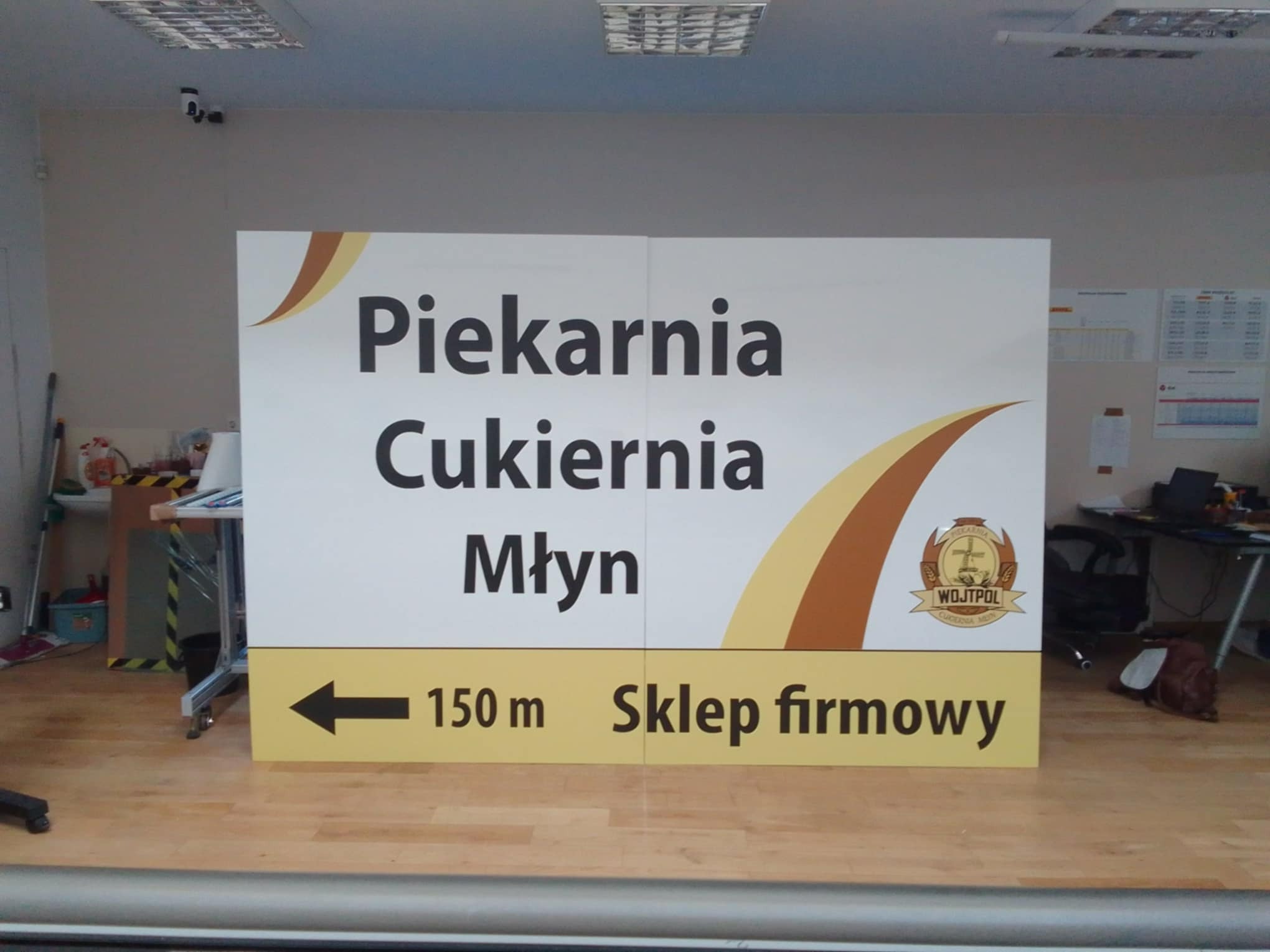 Duży, wolnostojący szyld reklamowy z napisem 'Piekarnia Cukiernia Młyn' oraz strzałką wskazującą kierunek do sklepu firmowego, umieszczony wewnątrz pomieszczenia.