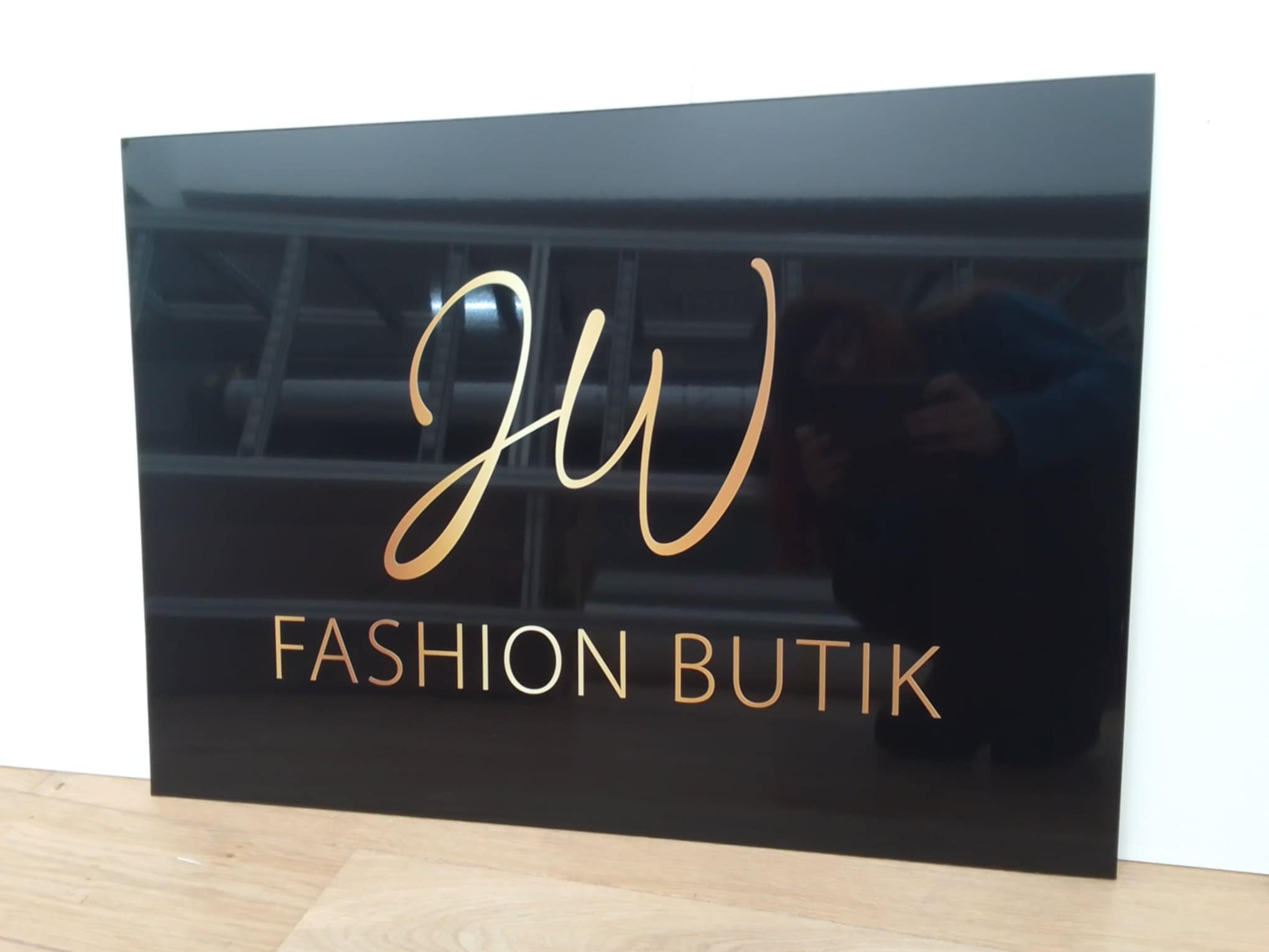 Czarna, błyszcząca tablica z napisem 'JW FASHION BUTIK' w kolorze złotym, ustawiona na drewnianej podłodze przy białej ścianie; widoczne odbicie osoby fotografującej.