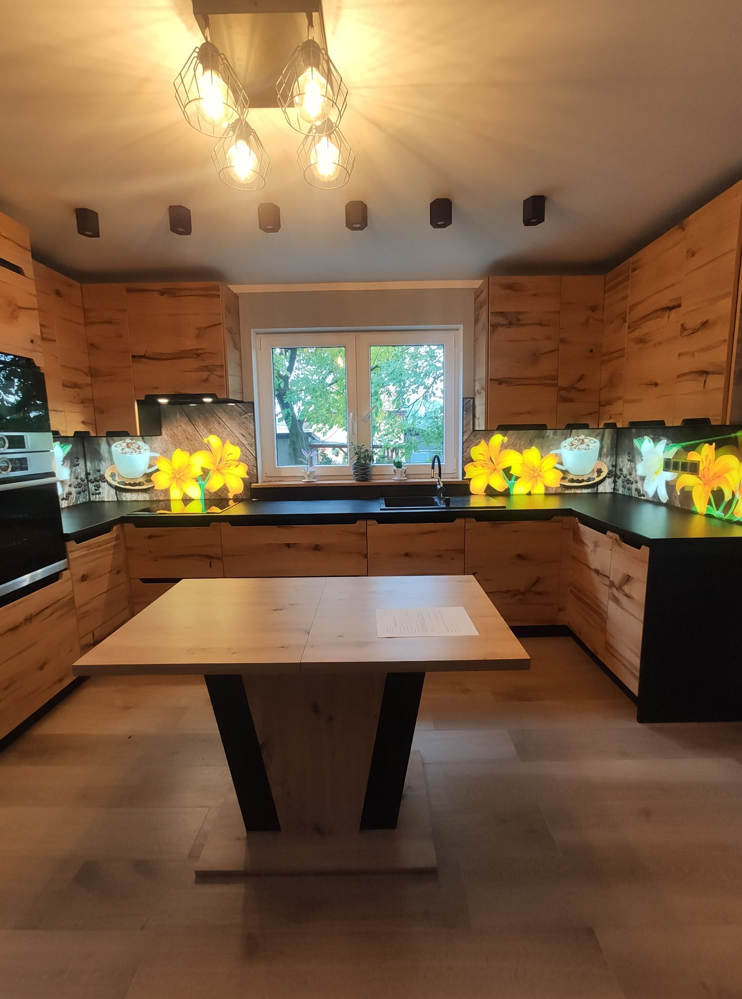 Nowoczesna kuchnia z drewnianymi frontami i czarnym blatem, ozdobiona panelem szklanym z motywem kwiatów i kawy, oświetlona wiszącą lampą z żarówkami Edison