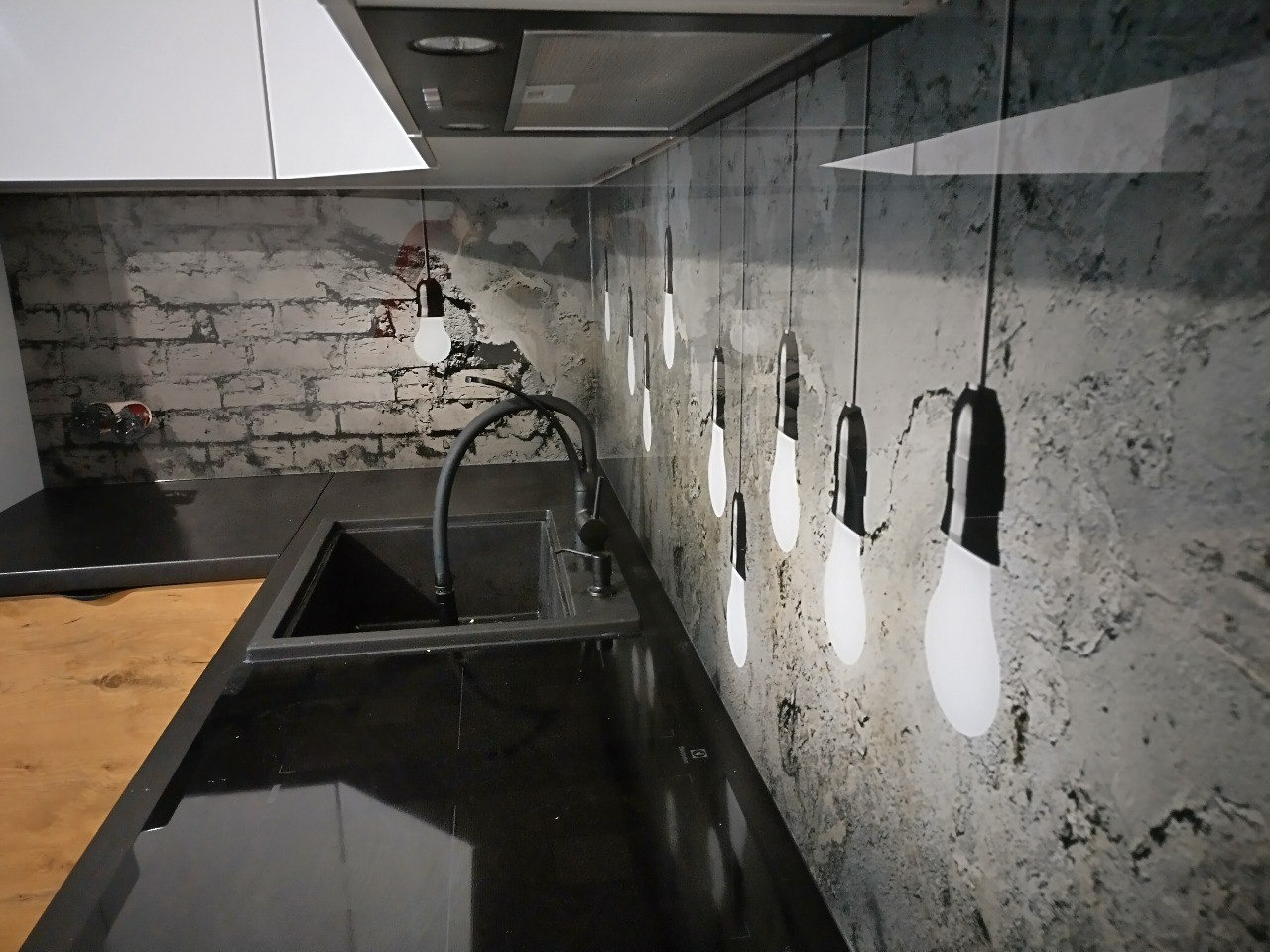 Czarny, matowy zlew kuchenny z baterią na tle ściany z fototapetą imitującą cegłę i żarówki na kablach.