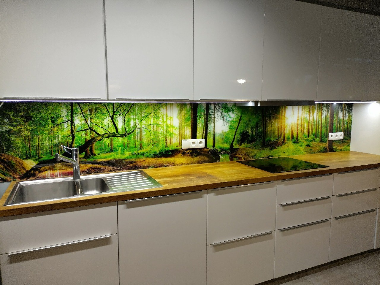 Szklana ściana nad blatem w kuchni z motywem leśnym, widoczny zlew, płyta grzewcza i białe szafki.