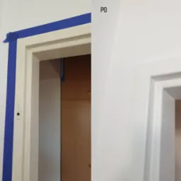 Porównanie stanu futryny drzwiowej przed i po odświeżeniu: po lewej stronie zabezpieczona niebieską taśmą malarską, po prawej świeżo pomalowana na biało, widoczne różnice w wykończeniu i czystości.