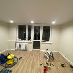 Twoja Ekipa Remontowa Konrad Lewicki - Jasne wnętrze po remoncie: świeżo pomalowane ściany, nowe panele podłogowe, okno balkonowe. Widoczne narzędzia i drabina malarska.