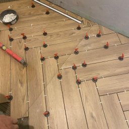 Twoja Ekipa Remontowa Konrad Lewicki - Układanie parkietu w jodełkę z użyciem systemu poziomowania płytek. Widoczny poziomica, wiadro z krzyżykami dystansowymi i fragment dłoni glazurnika.