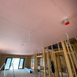 Twoja Ekipa Remontowa Konrad Lewicki - Montaż płyt karton-gips na suficie poddasza z widoczną konstrukcją drewnianą, instalacją i drabinami. Sufit w kolorze różowym.