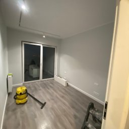 Twoja Ekipa Remontowa Konrad Lewicki - Pomalowane na szaro wnętrze pokoju z panelami, widoczny odkurzacz i drabina malarska. Świeżo wykończone ściany i podłoga po remoncie.